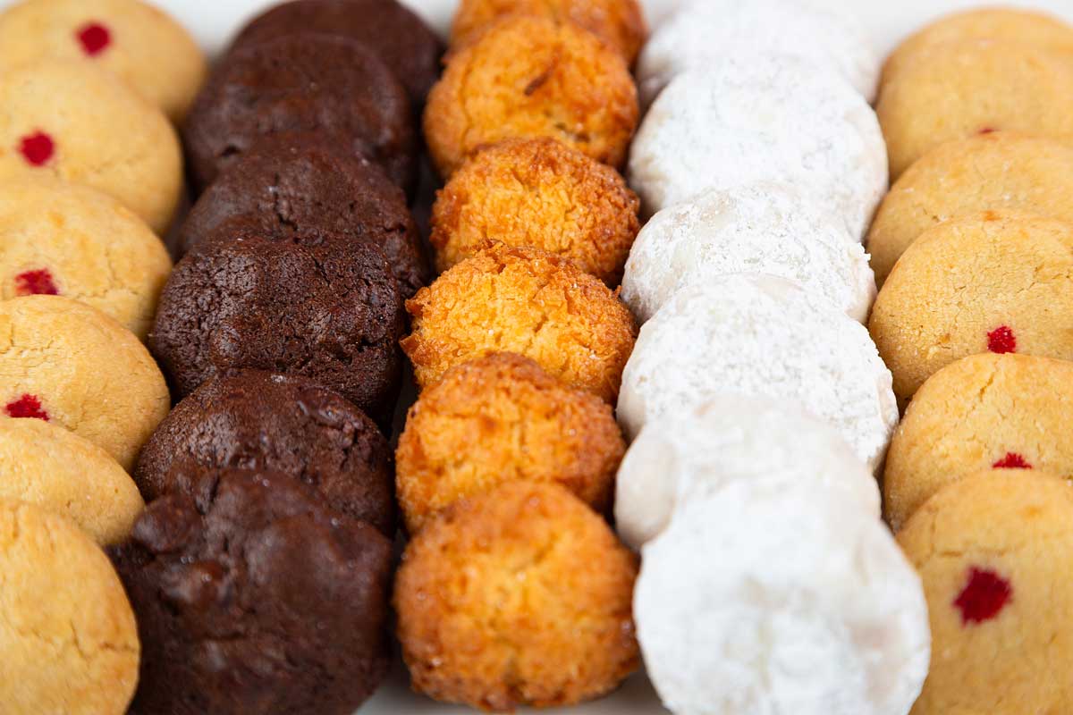 A close-up of an assortment of cookie dessert bites from a Maui bakery arranged in neat rows. The assortment includes chocolate cookies, coconut macaroons, powdered sugar-covered cookies, and vanilla cookies with a small red dot in the center. The cookies are displayed on a white surface.