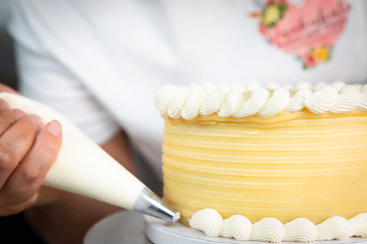 A person pipes white frosting onto the base of a yellow cake using a piping bag in a cozy Maui bakery. The cake is adorned with elegant horizontal lines and a wavy frosting border on top. The person is visible from the neck down, dressed in a pristine white shirt.