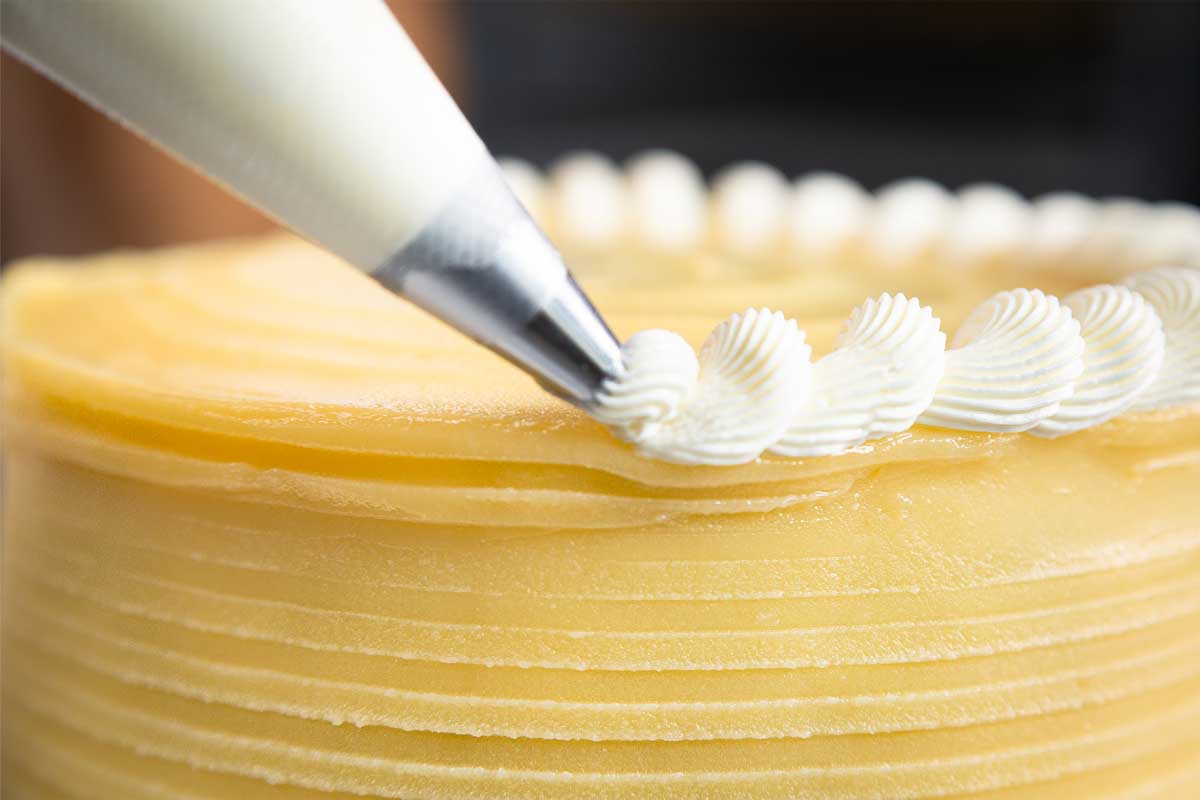 A close-up of a cake being decorated with white frosting using a piping bag at a Maui bakery. The cake has a smooth, textured yellow surface, and the piping bag is adding a delicate, wavy border to the edge. The background is blurred.