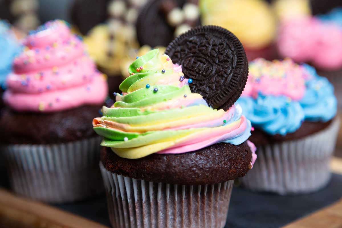 A close-up of a chocolate cupcake topped with brightly colored rainbow frosting and an Oreo cookie. The frosting, swirled with shades of green, yellow, purple, and pink, is adorned with colorful sprinkles. This delightful treat from a Maui bakery has more cupcakes blurred in the background.