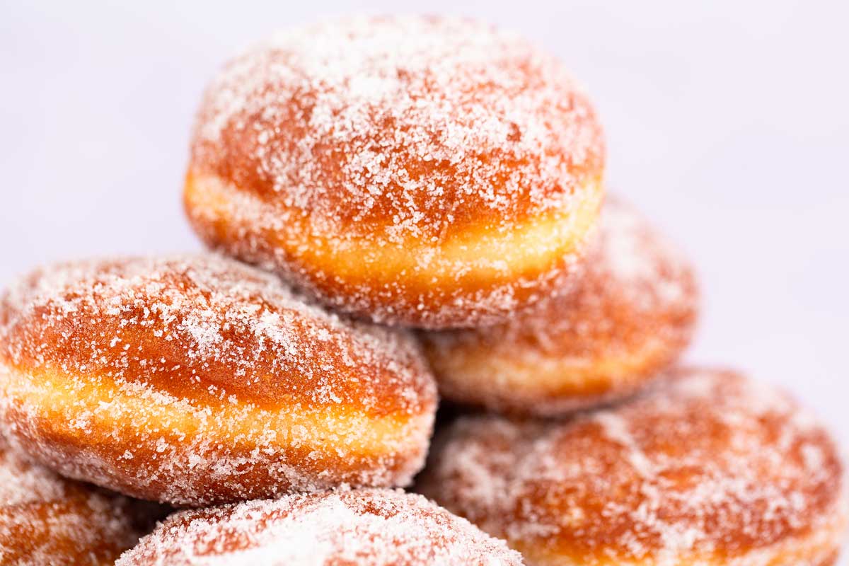 A close-up of several sugared malasadas stacked together, with a focus on their golden-brown exterior and the light dusting of sugar crystals glistening on the surface. The malasadas have a fluffy texture and inviting appearance, just like those from a charming Maui bakery, set against a plain background.