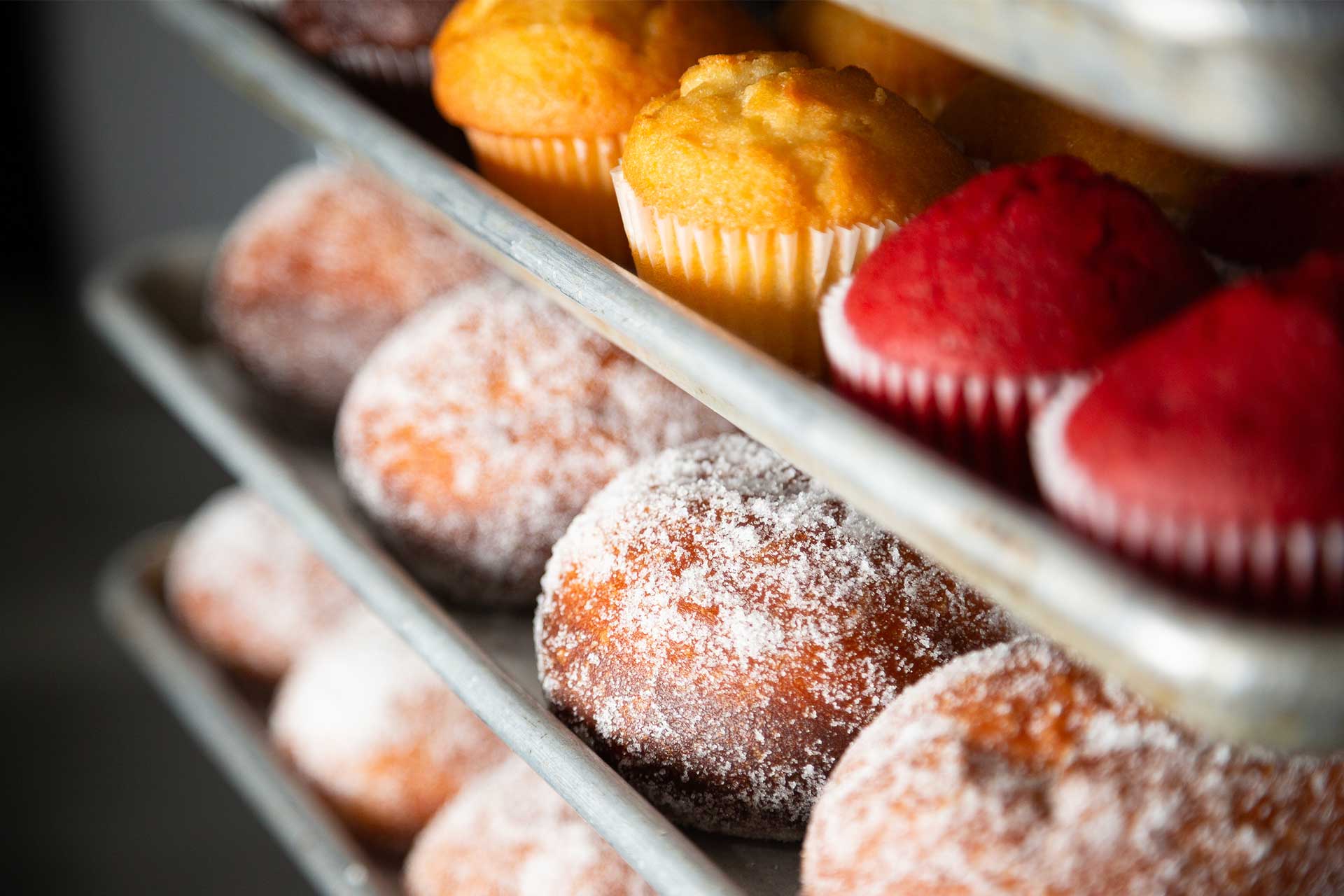 Close-up of baked goods on trays, including powdered sugar-covered malasadas, golden muffins, and red velvet muffins. The trays are stacked in a charming Maui bakery, showcasing a variety of freshly baked treats. The image exudes a warm, inviting atmosphere.