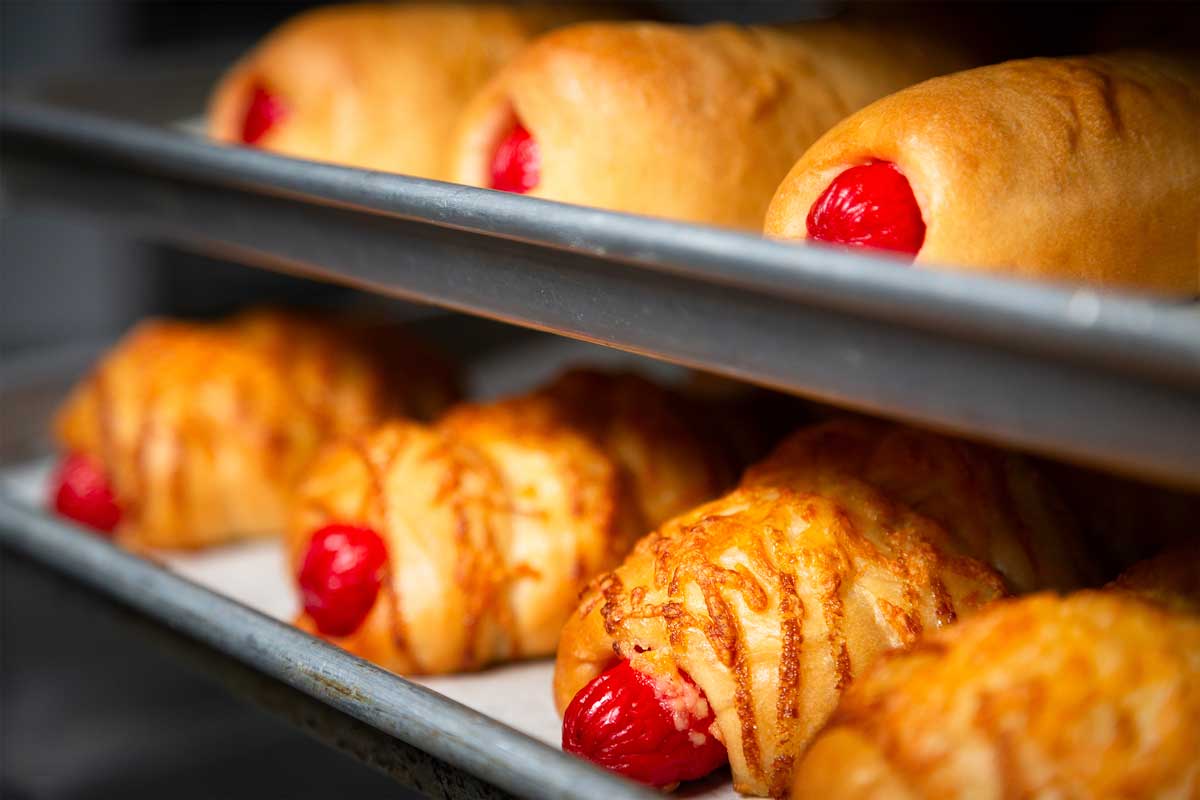 Close-up of several trays of freshly baked bread at a charming Maui bakery, wrapped around bright red sausages. The upper tray contains plain bread rolls, while the lower tray features rolls with melted cheese on top, giving a golden-brown appearance.