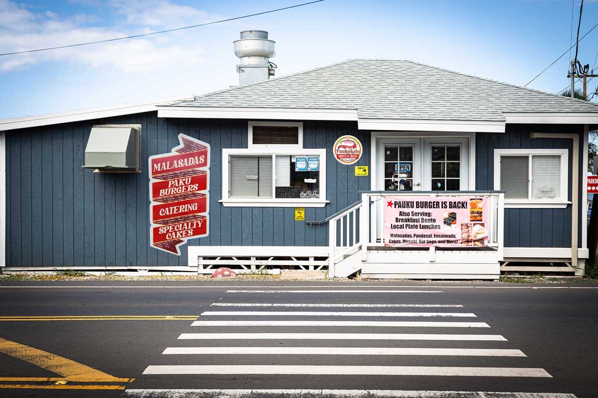 A small blue bakery with a white roof, located next to a crosswalk in Maui. Signs on the building advertise malasadas, Paku burgers, catering, and specialty cakes. A large banner on the right promotes Paku Burgers. The building has several windows and a white door.