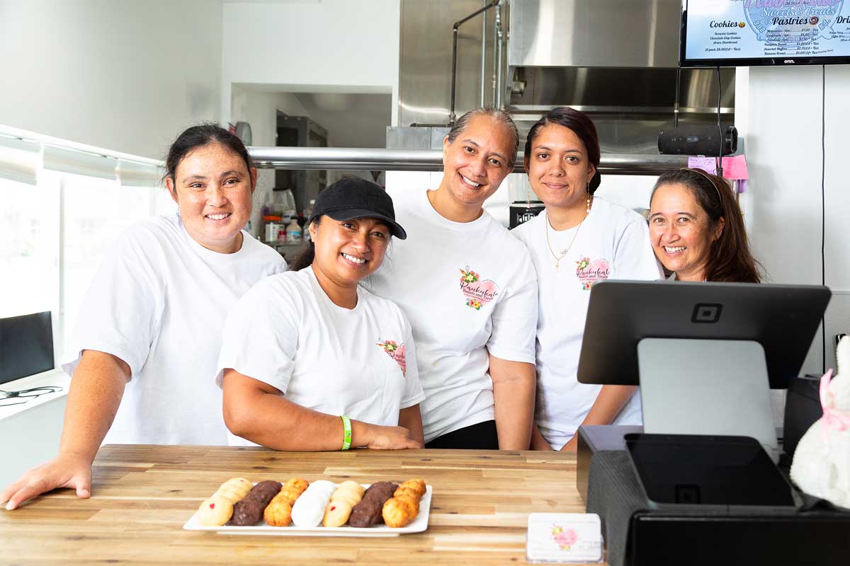 Five smiling women in white shirts stand behind a counter with a row of baked pastries in front of them. They are positioned in a sleek Maui bakery kitchen environment, with a digital register screen and menu visible behind them.