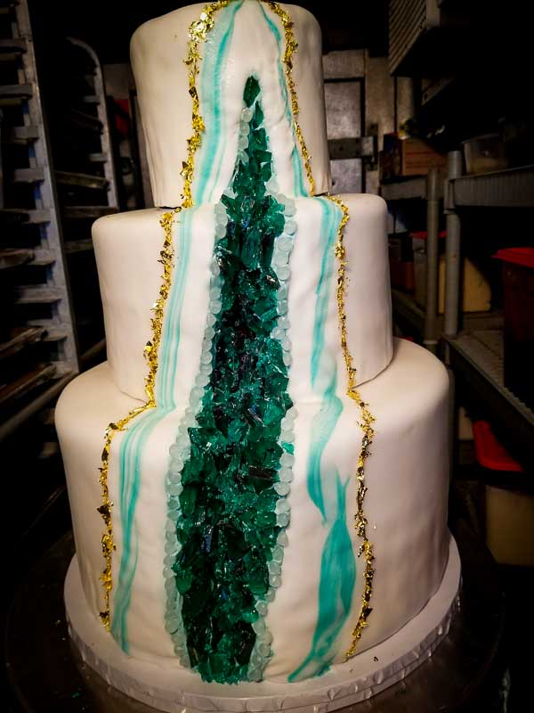 A three-tiered white cake with a geode design stands proudly in the bakery. It has a central vertical section featuring teal and green crystals with gold accents along the edges, mimicking a natural geode. The background is a cozy kitchen on Maui, complete with shelves and baking equipment.
