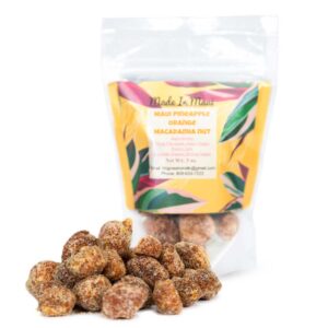 A clear plastic pouch labeled "Made in Maui" with "Maui Pineapple Orange Macadamia Nuts" text on a colorful background stands behind a small pile of sugared macadamia nuts, reminiscent of treats from a local Maui bakery. The nuts are covered in a crystalline sugar coating.