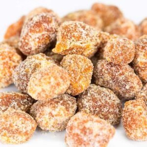 A close-up image of crystallized pieces of Maui Pineapple Orange Macadamia Nuts from a Maui bakery, coated in sugar. The small chunks are irregular in shape and have a rough, sugary texture. The colors range from light to dark amber, with the sugar crystals glistening under the light.