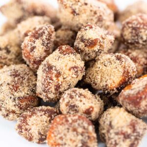 Close-up of a pile of Seasoned Cafe Macadamia Nuts from a Maui bakery. The nuts are irregularly shaped and covered in a rough, granular sugar coating, giving them a crunchy appearance. The white background emphasizes the texture and color of the delectable Maui treats.