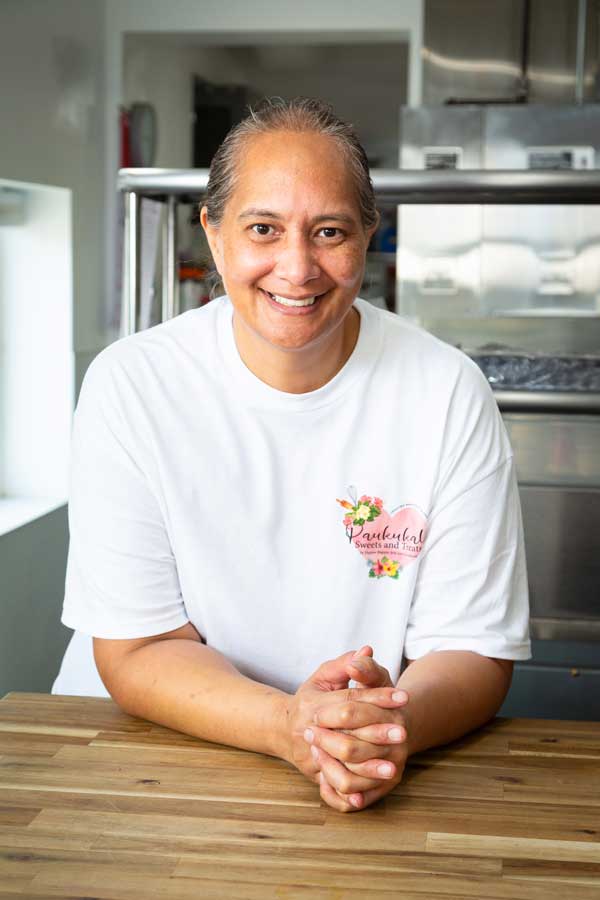 Chef Jocelyn Kanaele with a friendly smile stands behind a wooden counter in a kitchen, resting her hands on the surface. She is wearing a white T-shirt with a floral logo on the chest. In the background, reminiscent of a cozy Maui bakery, there are shelves and kitchen equipment.