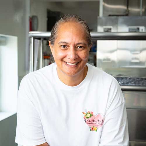 Chef Jocelyn Kanaele with a warm smile, wearing a white T-shirt with the logo "Pakūkalo Sweets and Treats," stands in a bakery kitchen. Commercial kitchen equipment is visible in the background, hinting at their delicious creations from Maui.